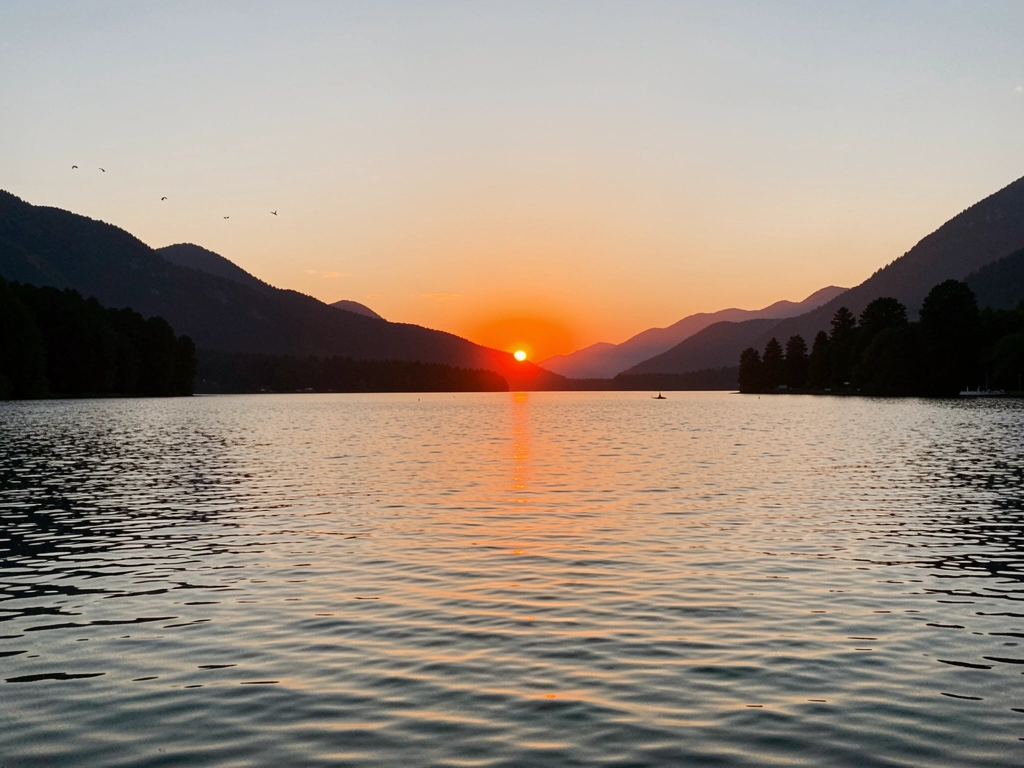Pôr do sol alaranjado sobre lago calmo com montanhas e árvores em silhueta, pássaros ao longe e reflexos suaves na água.