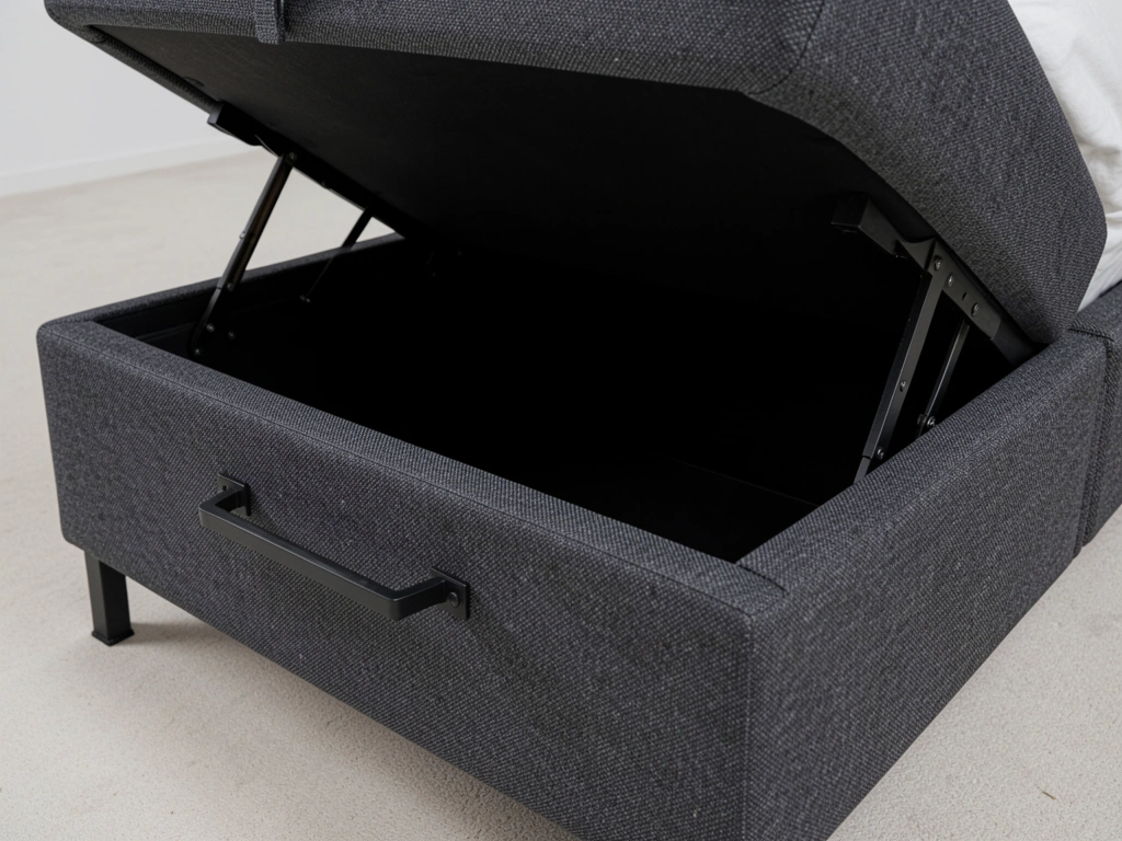 Cama estofada cinza escuro com base elevatória retangular aberta, forro preto, alças metálicas escuras e pés de metal, iluminação suave destacando a textura do tecido.