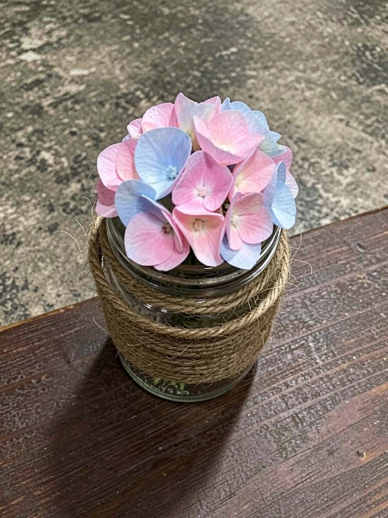 Pote de vidro envolto em barbante com uma flor natural dentro, sobre superfície de madeira.