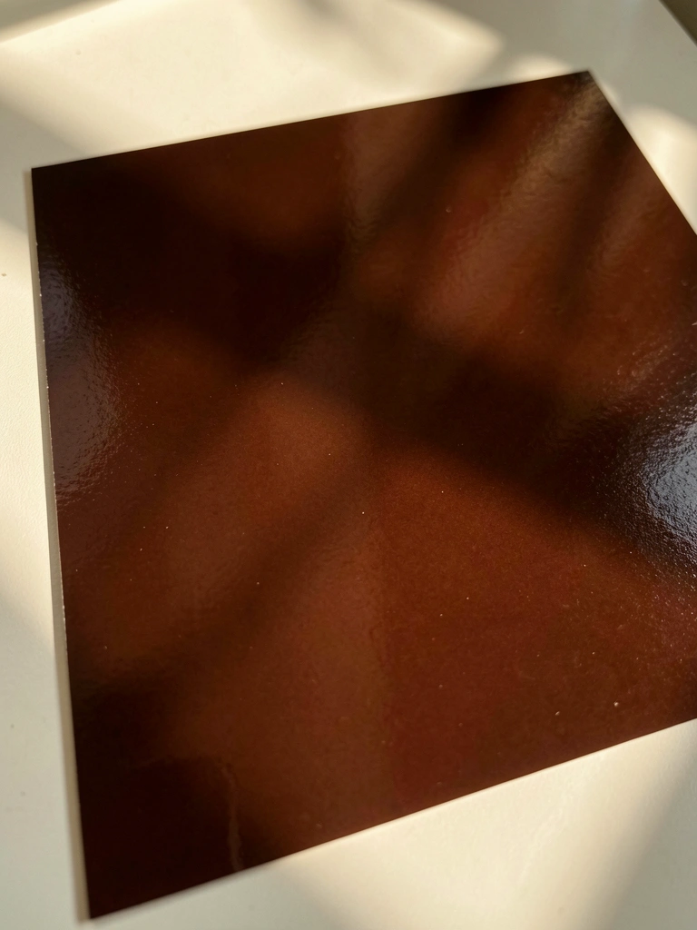 Amostra de tinta marrom escuro com superfície lisa e reflexos suaves sob luz natural, fundo off-white.