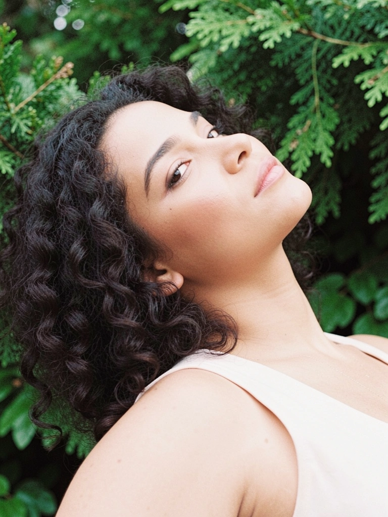 Mulher com cabelo cacheado escuro reclinada, folhagem verde ao fundo.