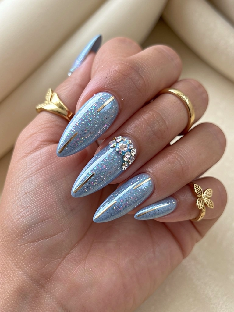 Mão com unha azul gel glitter, strass e linha dourada, anéis de ouro, luz natural difusa, fundo bege desfocado.