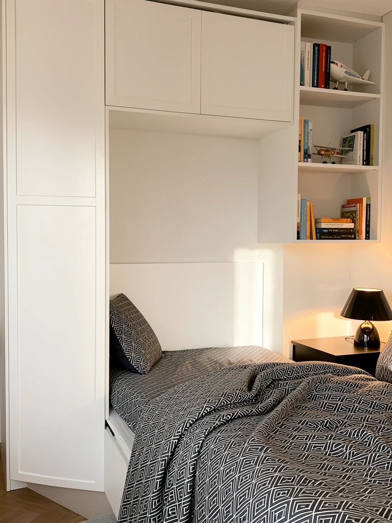 Luz natural incide sobre guarda-roupa modular branco com prateleiras, cama retrátil com edredom geométrico cinza e branco, mesa de cabeceira preta com abajur aceso e livros organizados.
