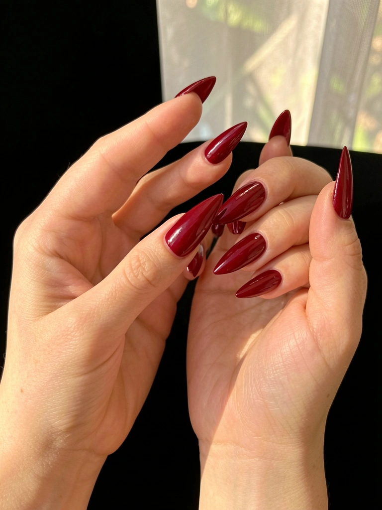 Close-up de mãos com unhas compridas e pontiagudas em esmalte vermelho cereja brilhante, fundo preto e luz natural difusa.