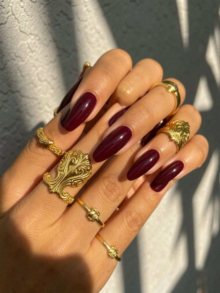 Mãos femininas com unhas vinho detalhadas e anéis dourados, luz natural difusa e fundo de parede texturizada cinza.