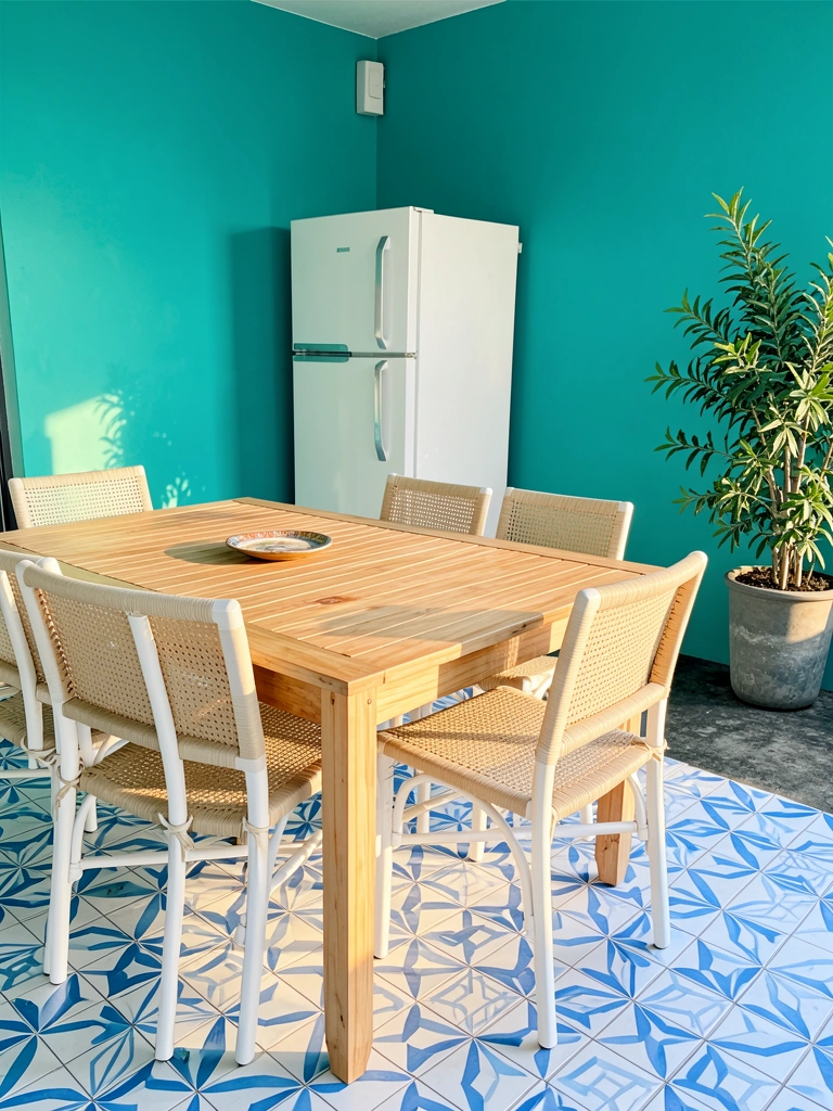 Mesa de madeira clara com cadeiras de vime brancas, parede azul-petróleo e piso geométrico azul e branco, luz natural.