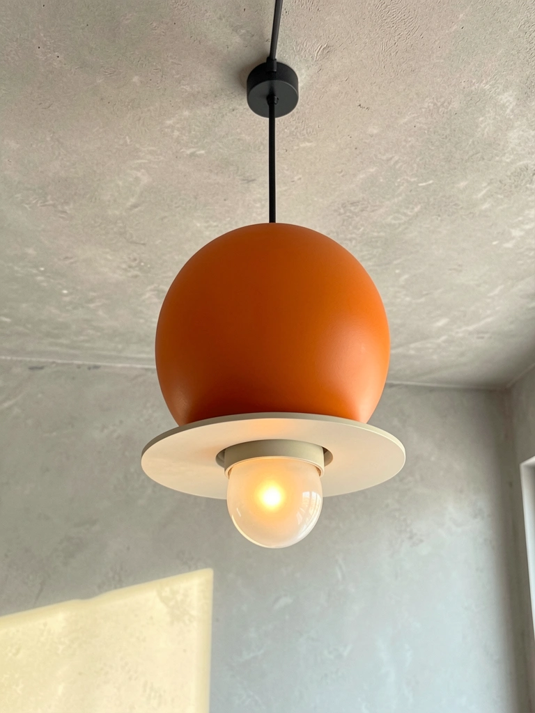 Luminária pendente com cúpula esférica laranja e difusor circular branco opaco, fio preto, teto de concreto cinza claro texturizado, luz natural.