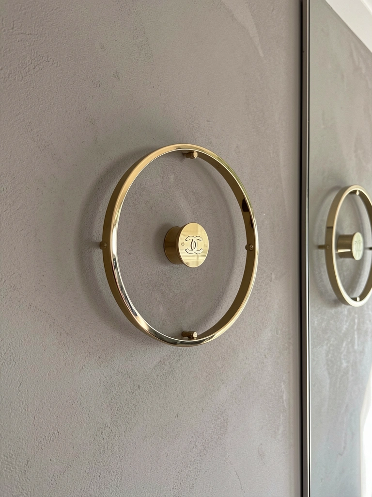 Lustre circular minimalista com aro dourado e placa Chanel, montado rente a parede texturizada cinza-rosada, refletido em superfície espelhada.