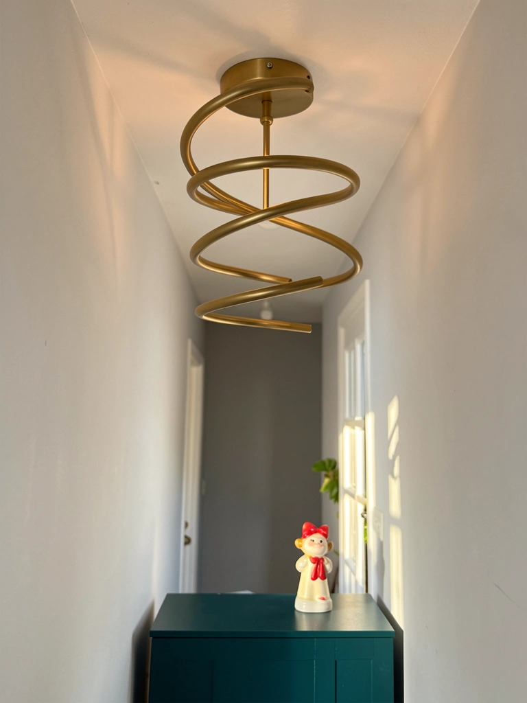 Luminária de teto espiral dourada minimalista em hall com parede cinza claro, armário verde-escuro e pequeno objeto decorativo com laço vermelho, luz natural entrando pela janela.
