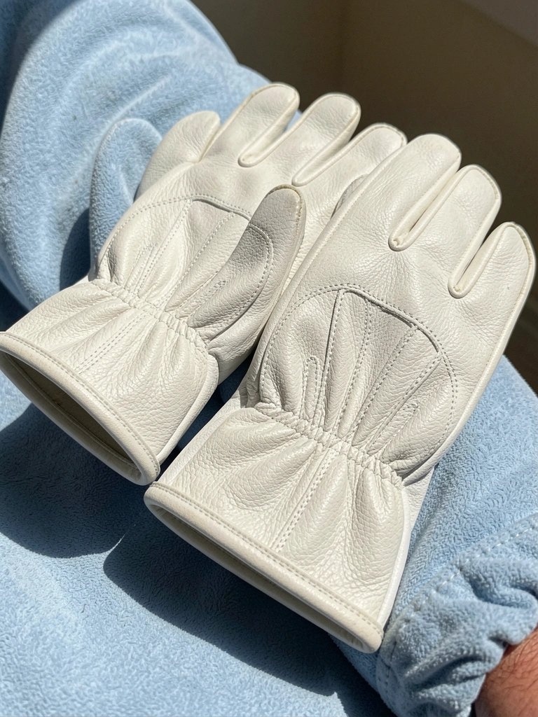 Luvas brancas de couro com costura reforçada sobre manga azul-clara, luz natural destacando a textura.