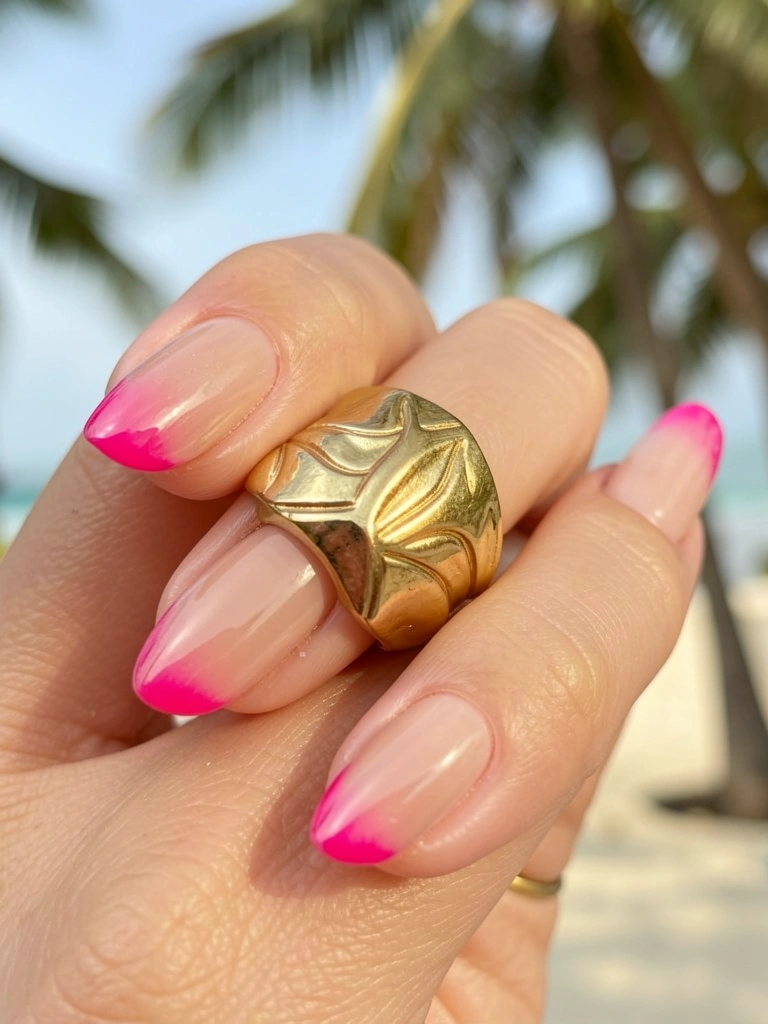 Anel dourado texturizado sobre unha nude com ponta rosa choque, luz natural difusa e fundo desfocado tropical.
