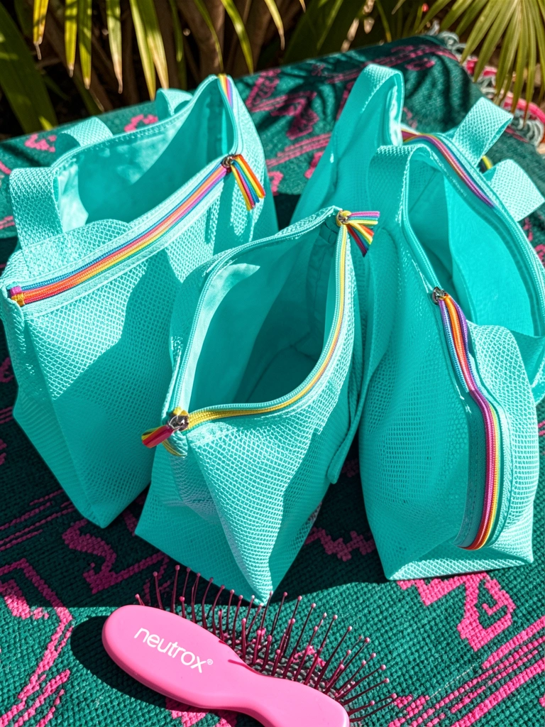 Três bolsas turquesa de nylon com zíperes arco-íris sobre tecido geométrico azul-petróleo e rosa, escova rosa ao fundo, luz natural.
