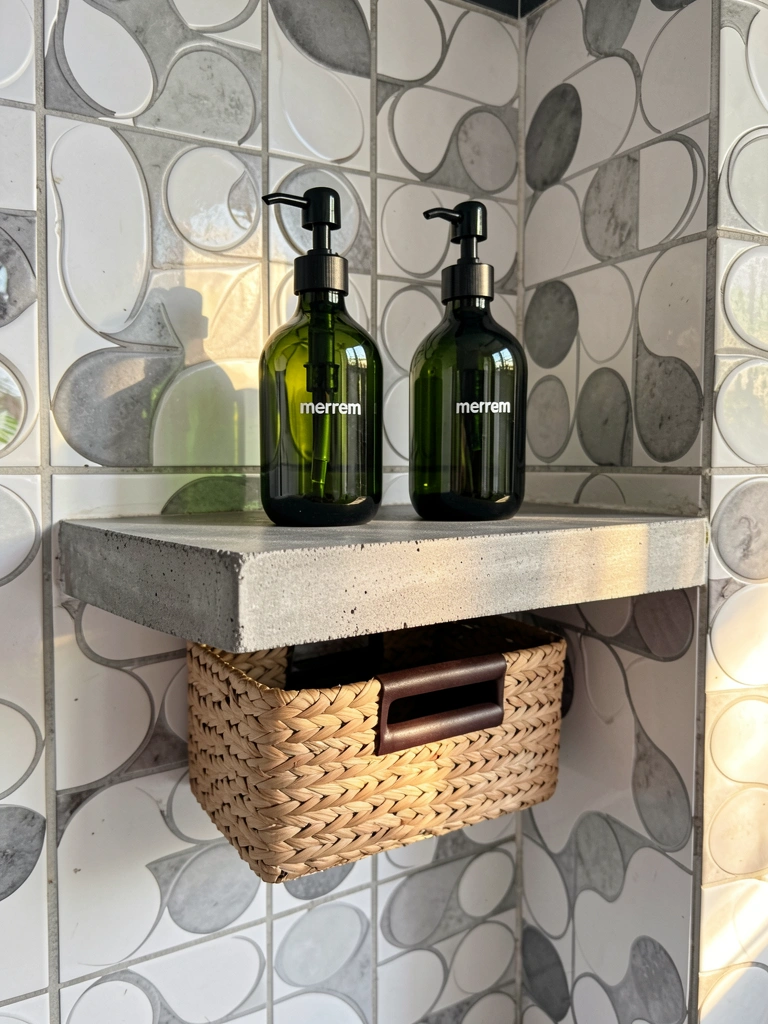 Prateleira de concreto cinza com dois frascos verdes escuros e cesta de vime, luz natural em banheiro com azulejos circulares.