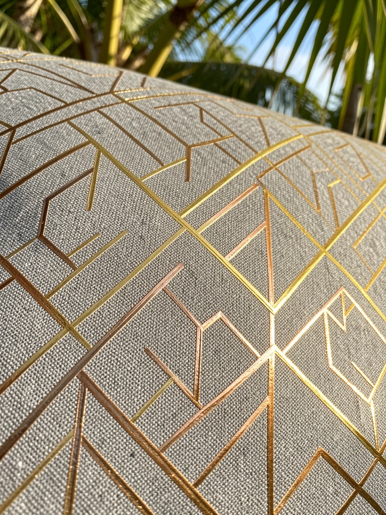 Tecido cinza texturizado com padrão geométrico de losangos e quadrados entrelaçados em linhas douradas e bronze, luz natural do dia.