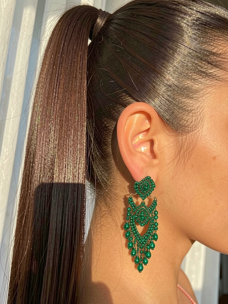 Cabelo escuro preso em rabo de cavalo alto com brincos verdes esmeralda de miçangas, luz natural e cortinas brancas ao fundo.