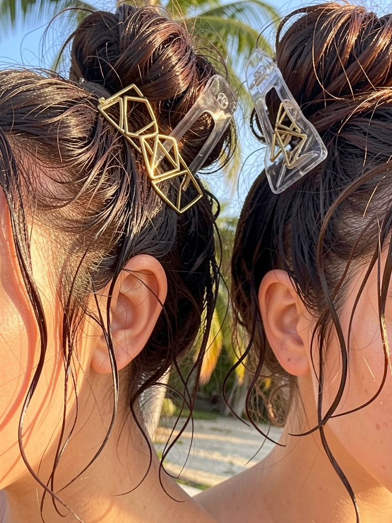 Duas mulheres com cabelo castanho escuro preso em coque bagunçado, com presilha geométrica dourada e presilha de acrílico transparente, luz natural quente e fundo com brisa tropical.