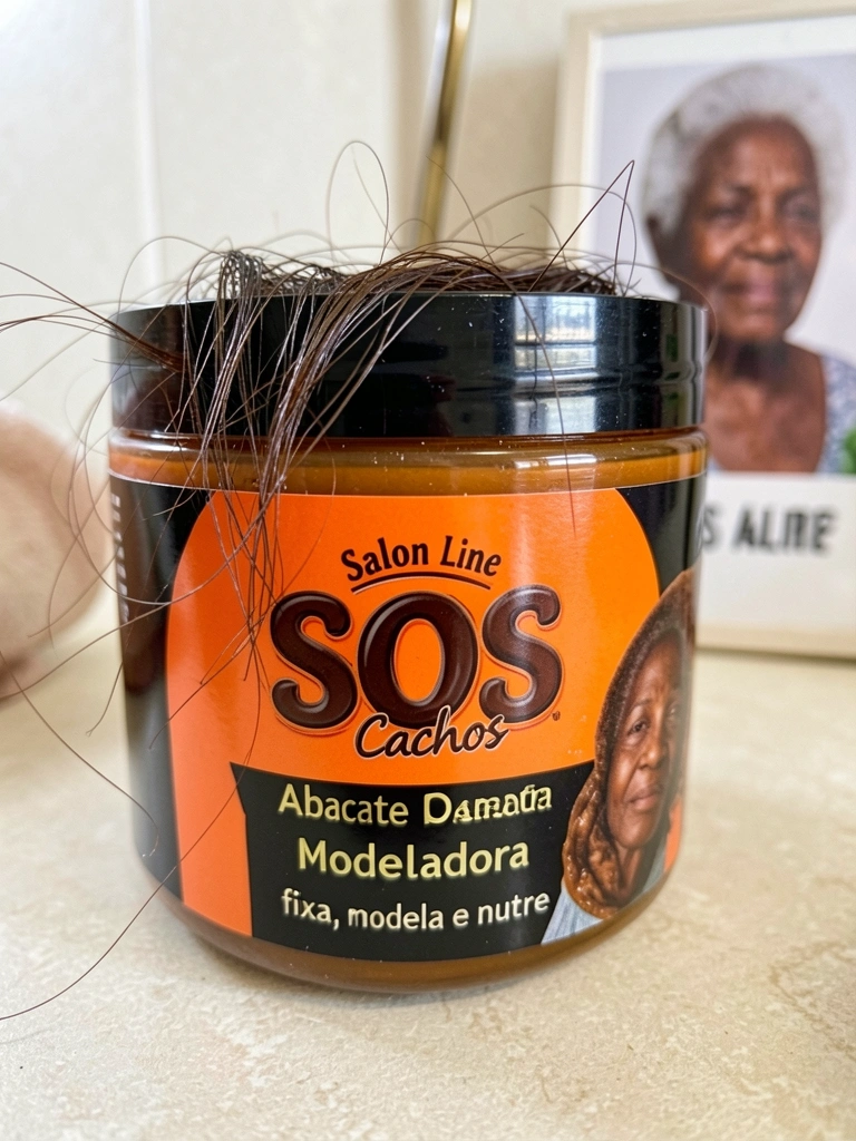 Pote de pomada modeladora Salon Line SOS Abacate, embalagem laranja e preta, com fios de cabelo sendo moldados; luz suave reflete na superfície; fundo desfocado com idosa negra.