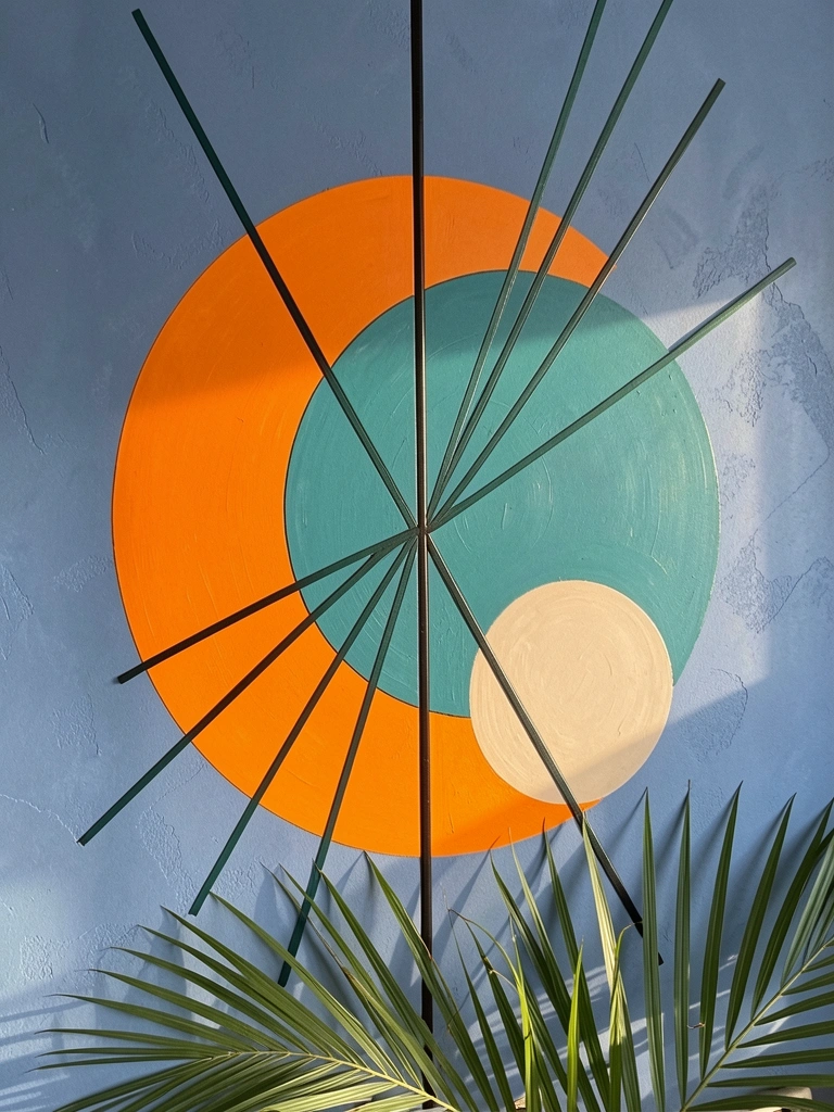 Arte abstrata com círculo laranja, teal e bege, linhas pretas e verdes, folhas de palmeira na base, fundo azul texturizado, luz natural.