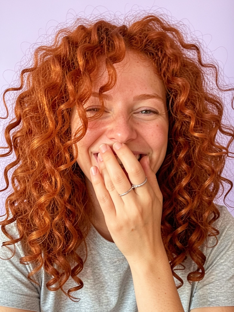 Mulher com cabelo cacheado ruivo vibrante, sorrindo com a mão na boca, camiseta cinza e anel prateado, fundo lavanda claro, luz suave destacando a textura dos cachos.
