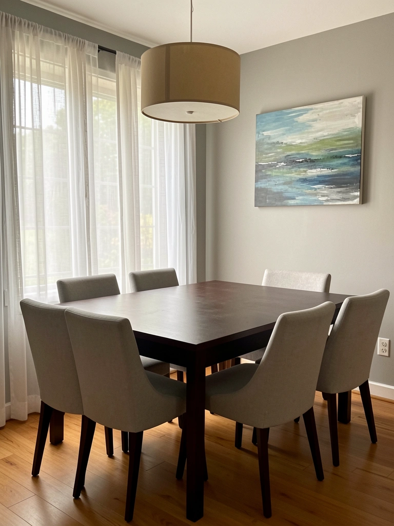 Mesa de jantar escura com seis cadeiras estofadas cinza claro, janela com cortinas brancas e pintura abstrata azul e verde na parede.