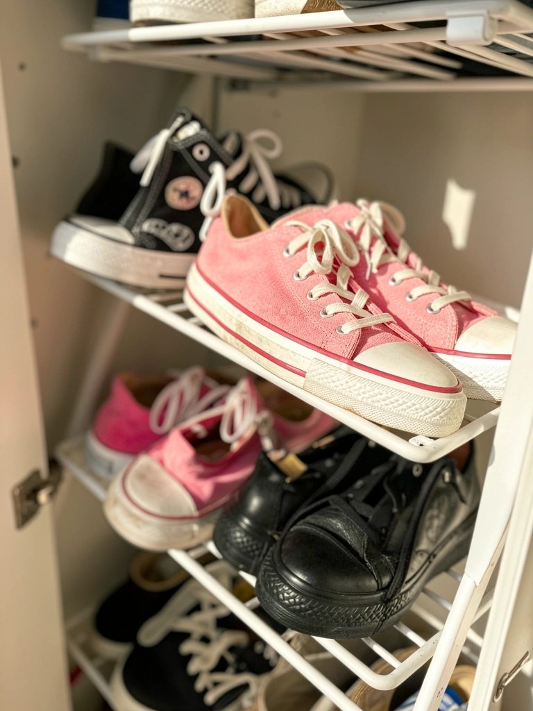 Sapateira de metal branco com tênis Converse preto, rosa camurça e couro preto, luz natural destacando cadarços e solados, fundo levemente desfocado.