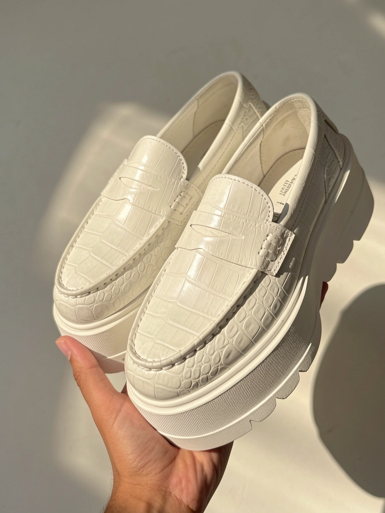 Mocassim branco com textura de crocodilo, solado plataforma, segurado por mão sobre fundo cinza claro com luz natural difusa.