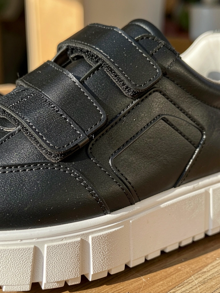 Tênis preto de couro com três tiras de velcro, sola branca grossa e painel geométrico, luz natural e fundo levemente desfocado.