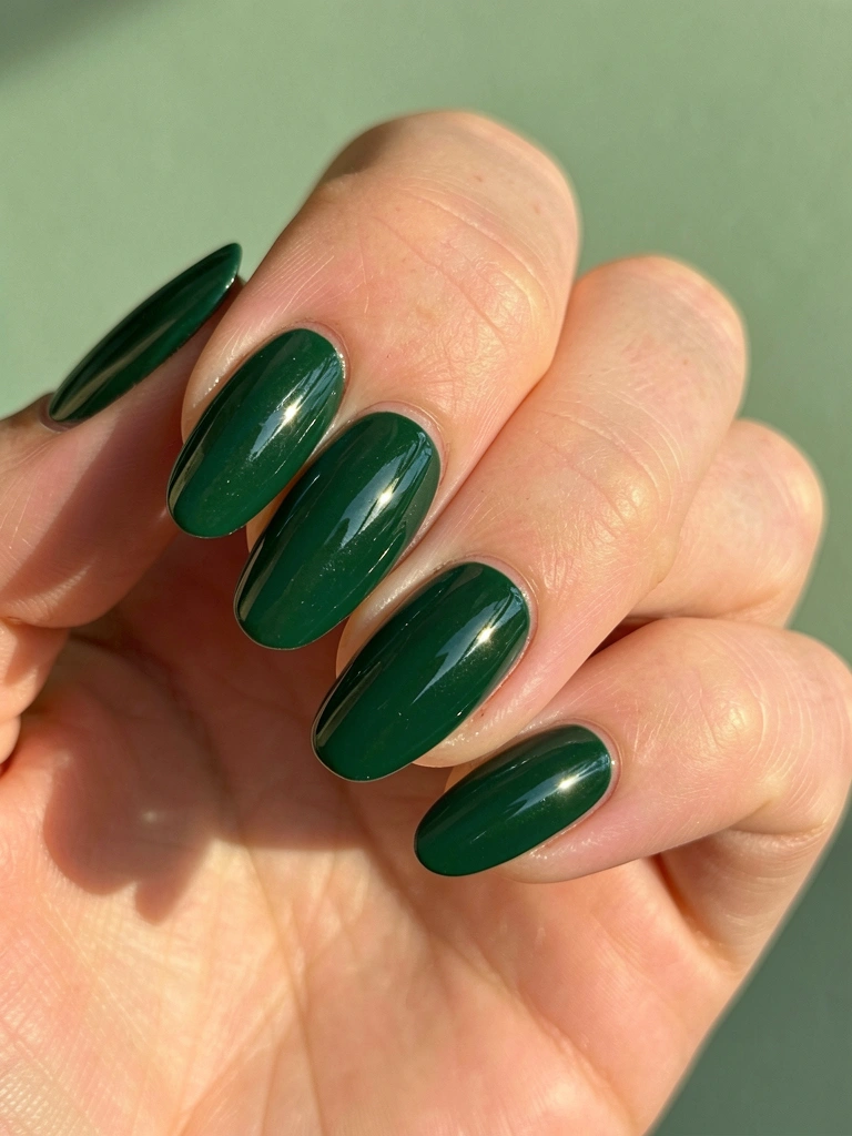 Mão feminina com unhas compridas e esmalte verde-esmeralda brilhante, reflexos de luz na superfície, fundo verde sálvia suave.