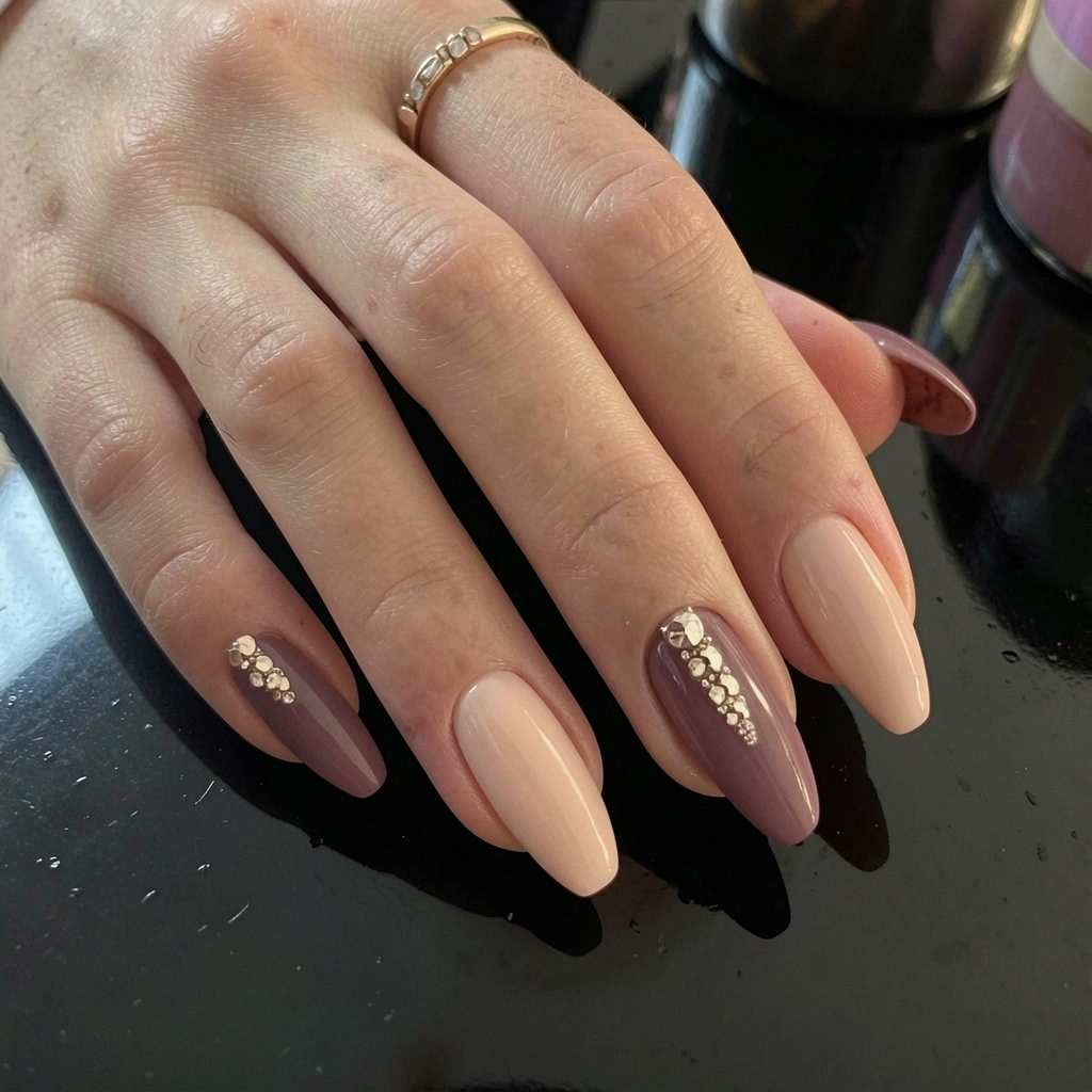 Mãos com unhas decoradas em esmalte nude e vinho escuro com pedrarias, apoiadas sobre superfície escura e reflexiva, transmitindo elegância e autocuidado.