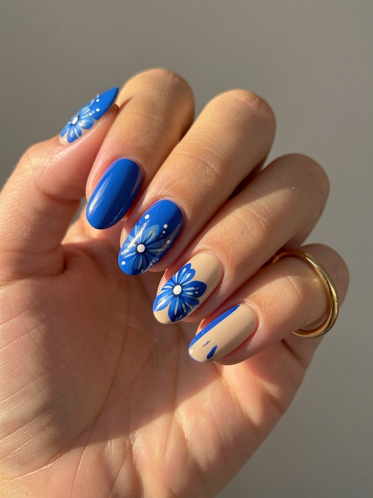 Mão feminina com unhas azuis e nude decoradas com florais brancos, anel dourado, luz natural suave e fundo cinza neutro.