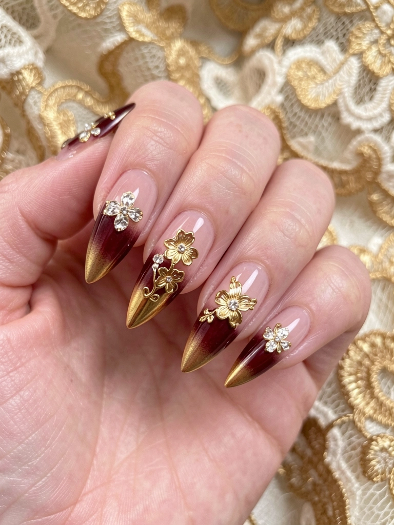 Mão com unhas em gel alongadas em gradiente de vinho a dourado com flores douradas e cristais, fundo de renda vintage desfocado.