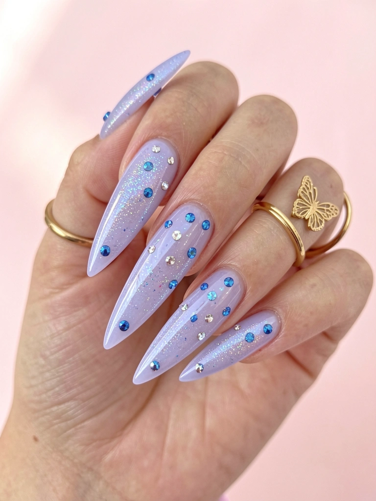 Mão com unhas alongadas em gel lavanda com glitter iridescente, pedras azuis e pratas, anéis delicados de ouro e fundo rosa suave.