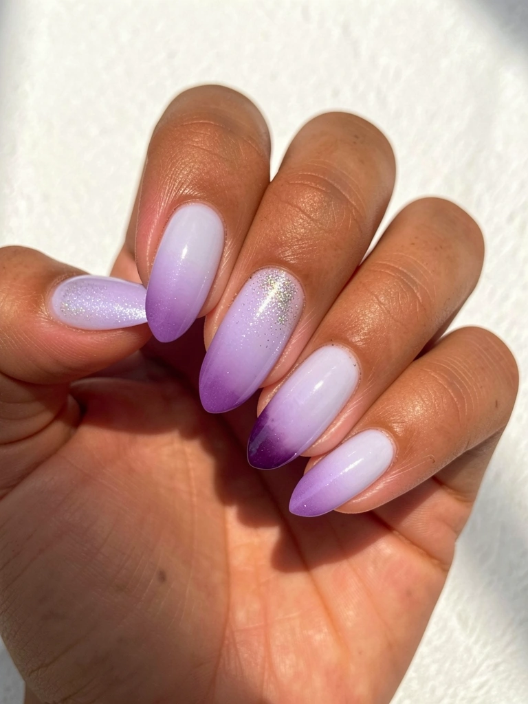 Mão com unhas ombré lavanda, do tom claro na base ao roxo escuro nas pontas, glitter sutil em uma unha, luz solar suave sobre fundo branco texturizado.