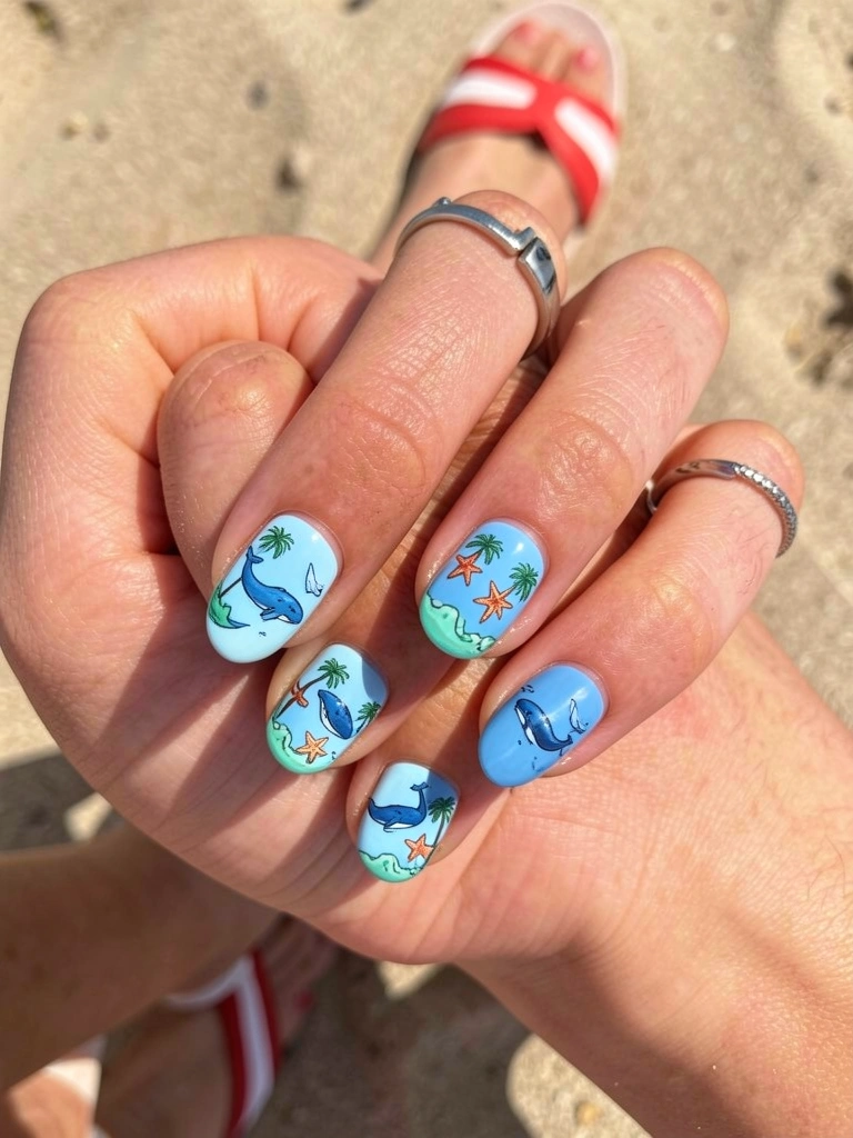Mãos com unhas azul-claro decoradas com baleias, estrelas-do-mar e palmeiras, anel de prata em um dedo, chinelo listrado ao fundo desfocado, luz natural.