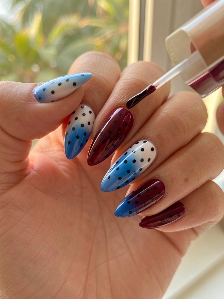 Mão aplicando esmalte em gel em unha longa com gradiente bordô a azul e bolinhas pretas, luz natural suave e folhagem ao fundo.