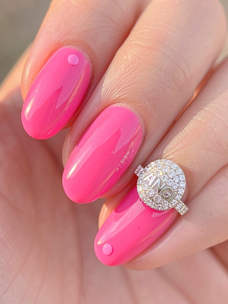 Mão com unha pintada de rosa brilhante e detalhe de bolinha, anel cravejado de diamantes com inscrição 