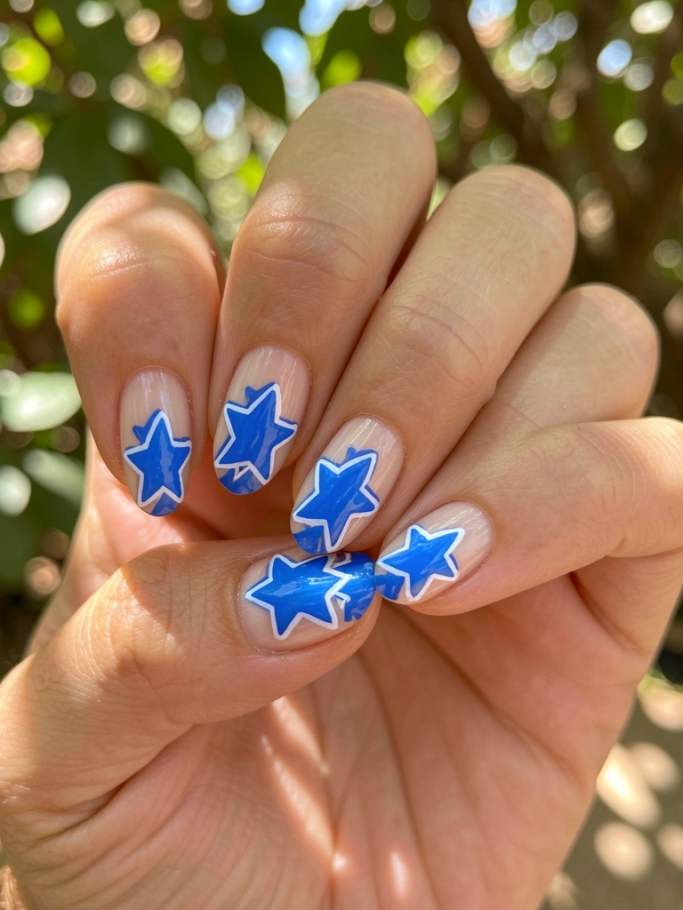 Mãos femininas com unhas naturais decoradas com estrelas azuis detalhadas em branco, luz solar filtrada por folhas ao fundo.