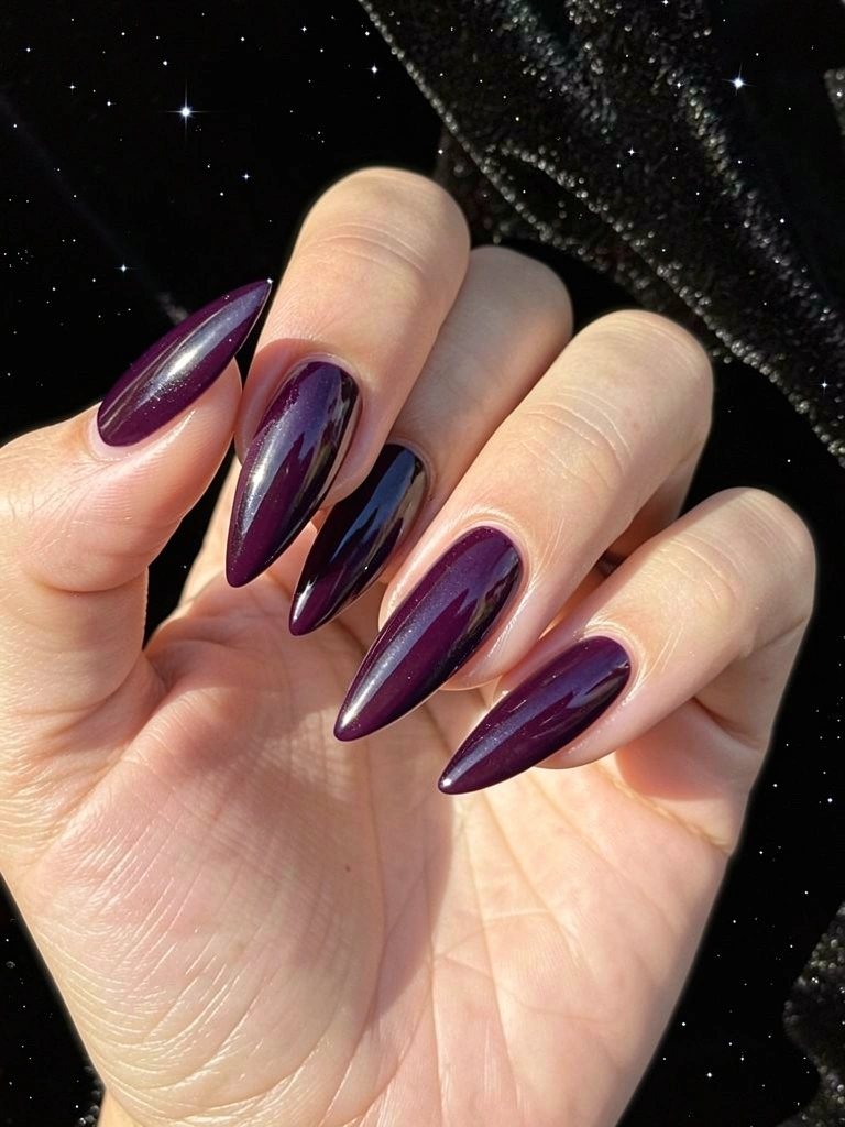 Close-up de mão com unhas alongadas e pontiagudas em esmalte roxo escuro cintilante, fundo preto aveludado com estrelas sutis.