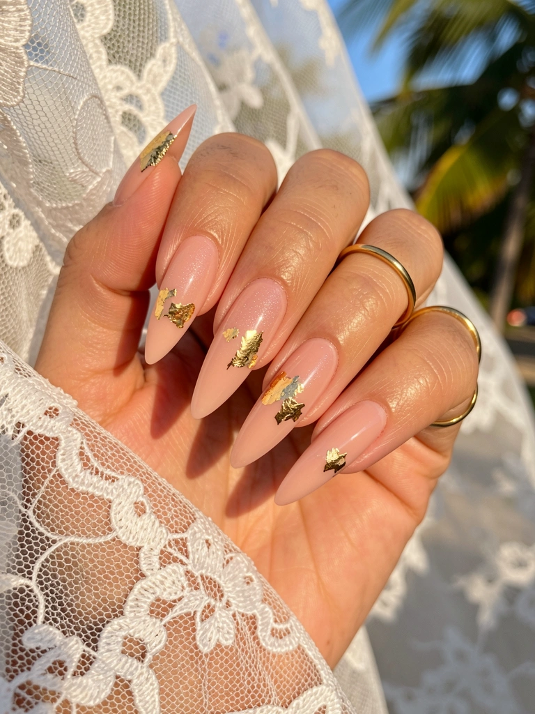 Mão feminina com unhas alongadas em gel nude rosado, detalhes em folha de ouro e anel fino, fundo de renda branca iluminado por luz natural.