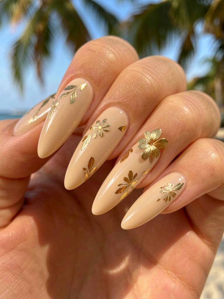 Close-up de mão com unhas longas em esmalte nude bege, detalhes em folha de ouro e florzinha dourada, luz natural e fundo desfocado.