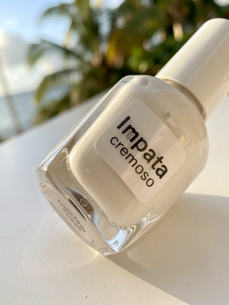 Frasco branco de esmalte Impata deitado sobre superfície clara, com luz natural difusa destacando o rótulo e o brilho suave do vidro.