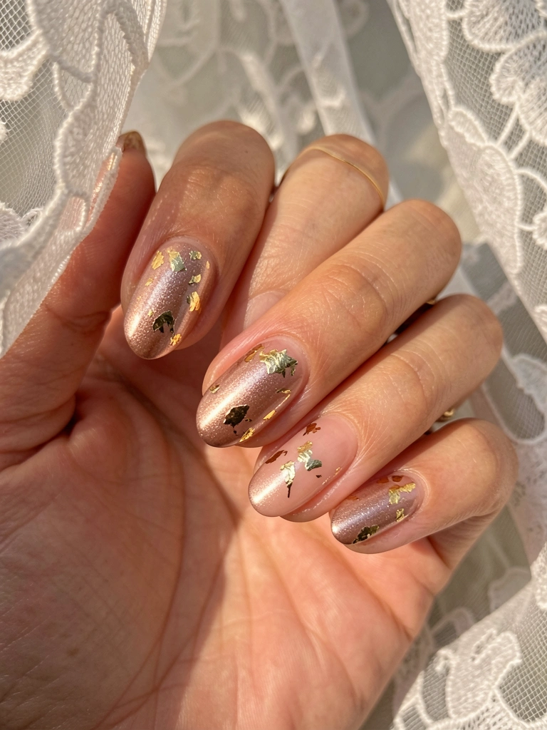Mão feminina com unhas nude, brilho rosé e flocos de ouro, anel fino e renda branca ao fundo, luz natural suave.