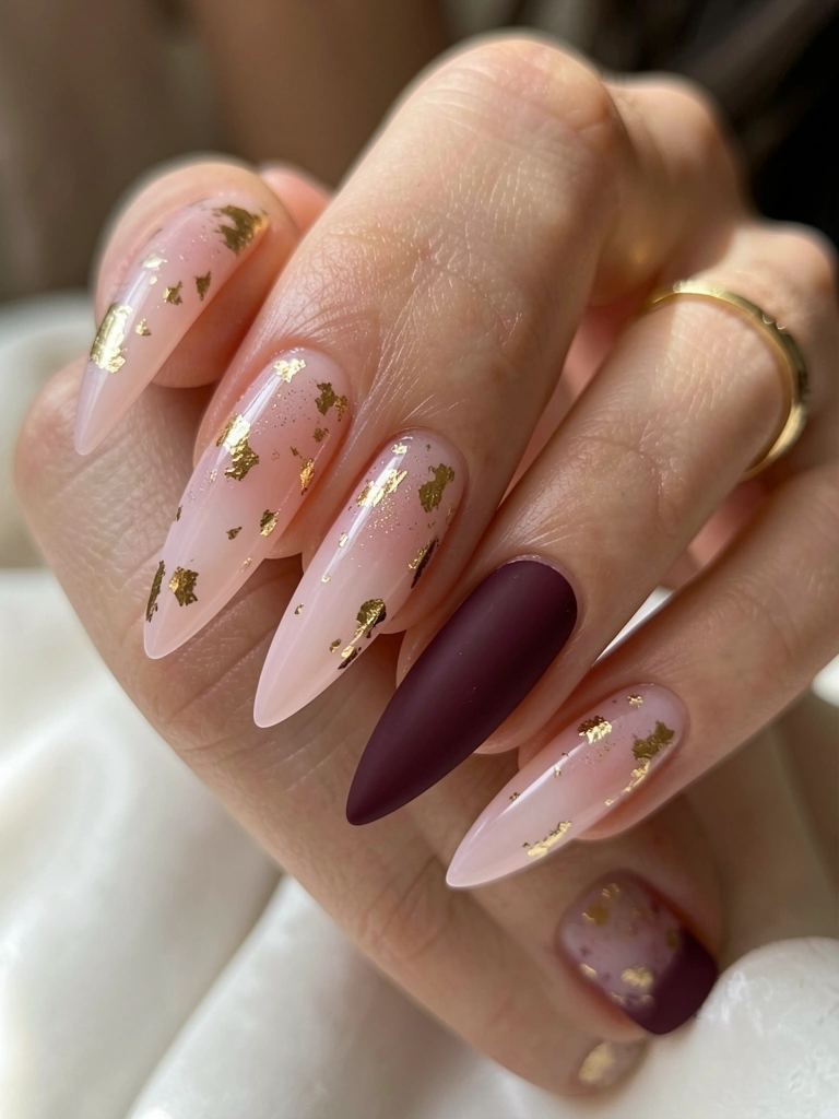 Mãos com unhas longas e pontiagudas, esmalte ombré rosa claro com folhas de ouro e uma unha vinho fosca, anel de ouro, luz natural suave.