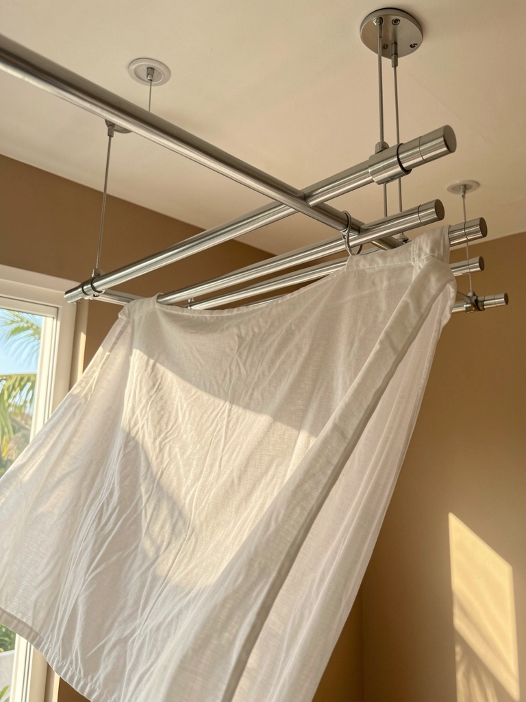 Sistema moderno de varal suspenso com cabides de prata e lençol branco esvoaçante, luz natural entrando por janela, sombras suaves no tecido translúcido.