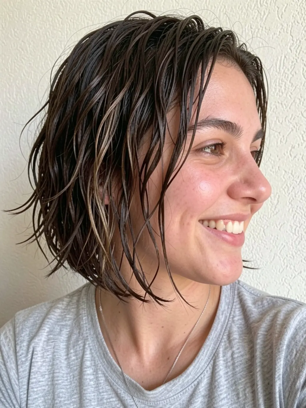 Perfil de mulher com cabelo castanho escuro em bob irregular, sorriso natural, luz suave de janela.
