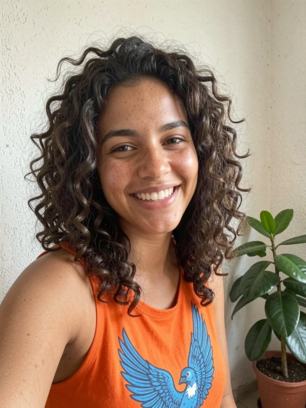 Mulher brasileira com cabelos castanhos escuros cacheados volumosos, sorrindo naturalmente, usando top laranja com estampa de pássaro azul, fundo com parede texturizada e vaso de planta.