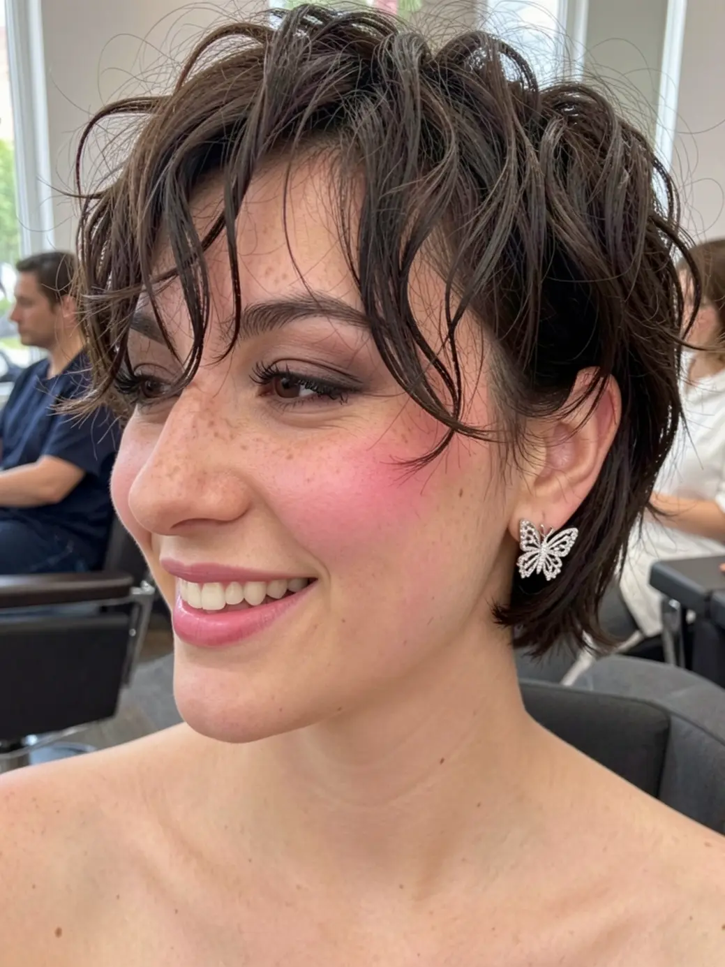 Jovem de cabelo cacheado curto e brinco de borboleta, sorrindo em salão com luz natural.