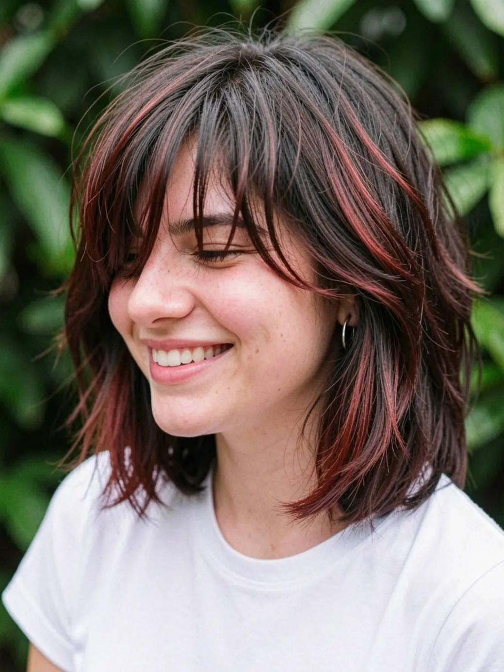 Jovem brasileira de cabelo castanho escuro com mechas avermelhadas, franja irregular e sorriso natural, usando camiseta branca e brincos de argola.