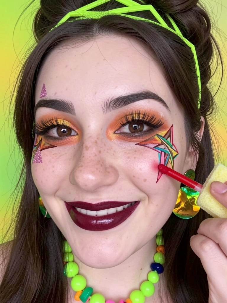Dedo aplicando batom líquido vermelho na bochecha, com esponja úmida ao lado.