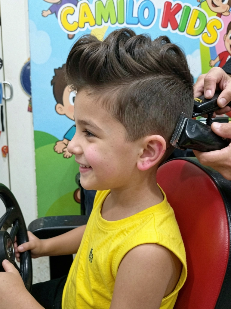 Menino sentado em cadeira de barbeiro com capa, cabelo com degradê lateral, barbeiro usando máquina.